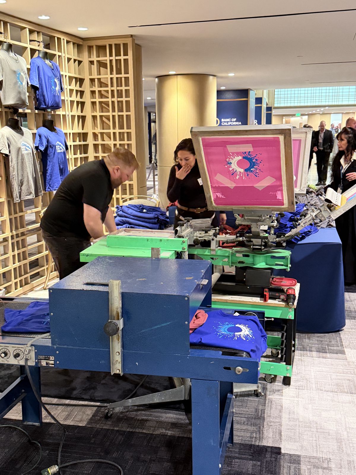 Press and conveyor dryer running inside a hotel event space with staff printing shirts